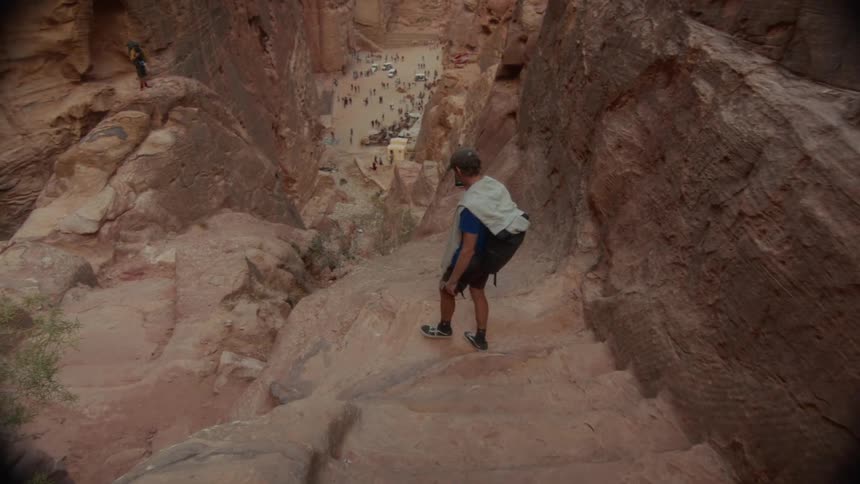 A lone hiker carefully descends a steep, ancient stone staircase carved into a red rock canyon, with a bustling courtyard visible far below
