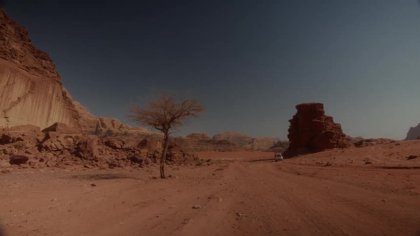 An off-road vehicle drives down a dusty path through a vast red desert landscape surrounded by rocky mountains