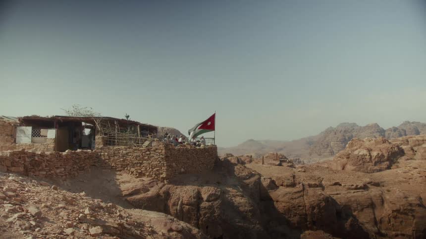 The Jordanian flag flies proudly over a rustic stone outpost perched on a cliff edge in the vast, rocky desert landscape