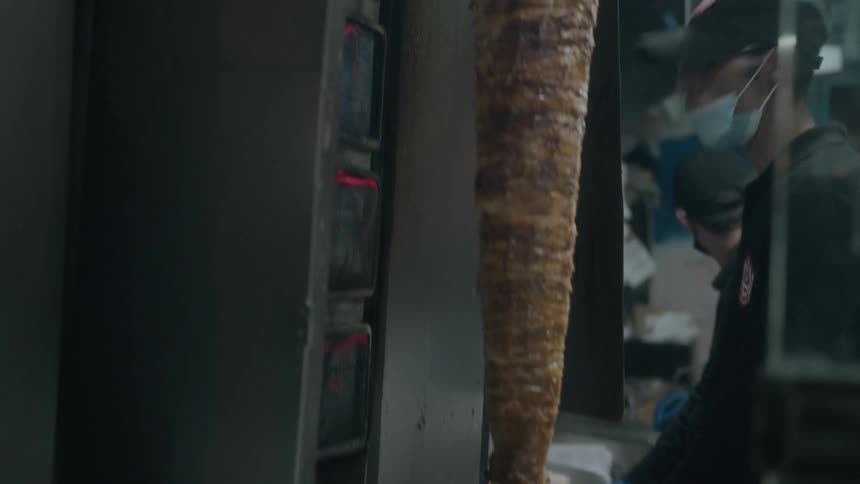 A vertical rotisserie of shawarma meat roasts in a bustling street food kitchen as chefs prepare orders in the background