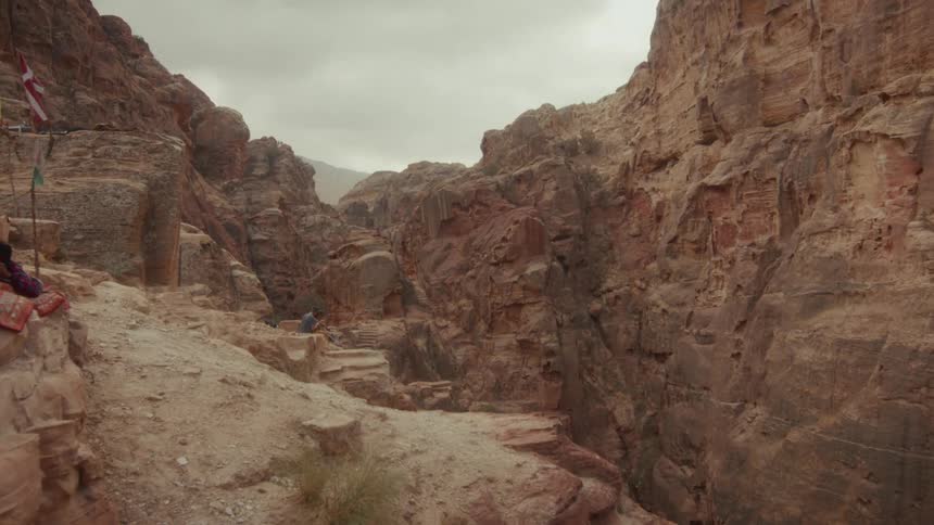 A lone person rests on ancient steps carved into the side of a vast, rocky canyon under an overcast sky