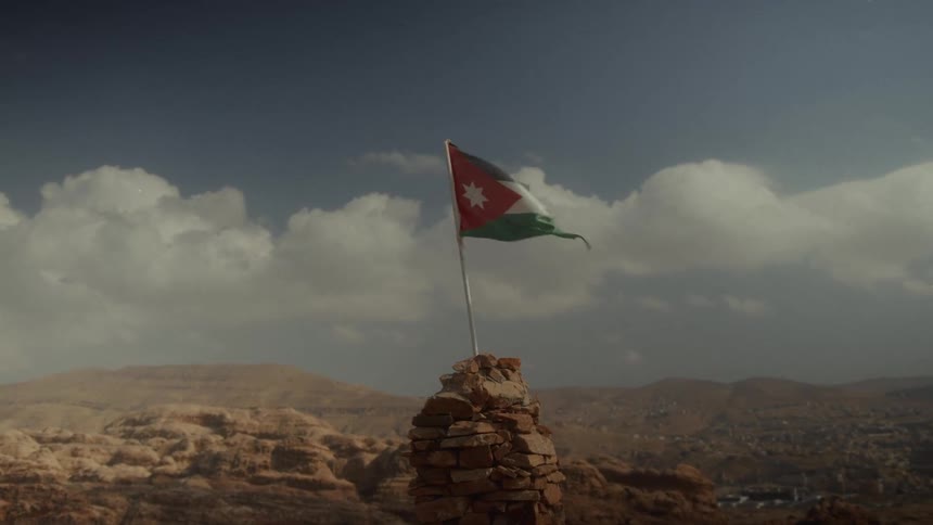 The flag of Jordan waves in the wind atop a stone pile, with a desert mountain landscape in the background