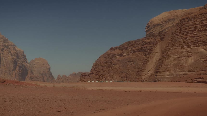 A static wide shot of a small camp at the base of a towering red sandstone mountain in the vast Wadi Rum desert