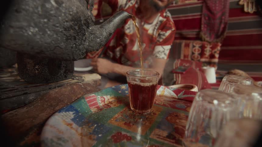 A man pours traditional hot steaming tea from a kettle into a glass in an authentic Bedouin tent in slow motion