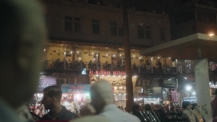 A bustling street market comes alive at night in a Middle Eastern city with a busy restaurant in the background