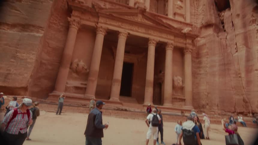 A wide, low-angle shot tilts down to reveal tourists admiring the ancient Al-Khazneh Treasury carved into the sandstone cliffs of Petra, Jordan