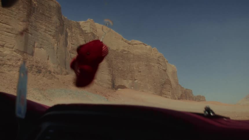A small red teddy bear keychain hangs and swings in a car driving through a vast desert landscape with large rock formations