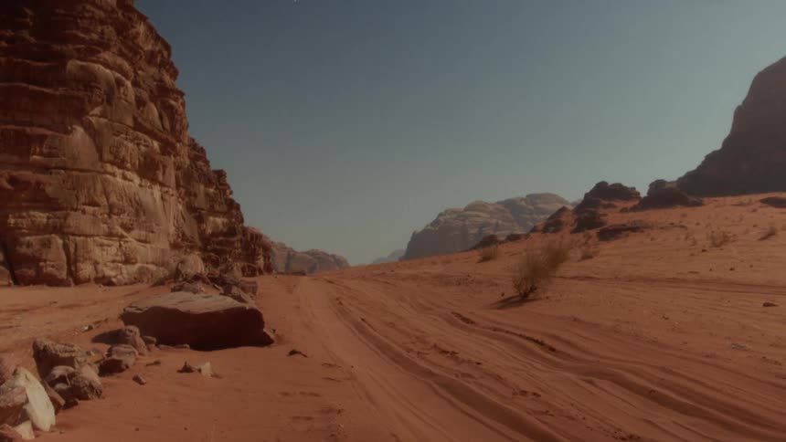 A static wide shot shows a sandy desert road winding through the red rock canyons of Wadi Rum, Jordan
