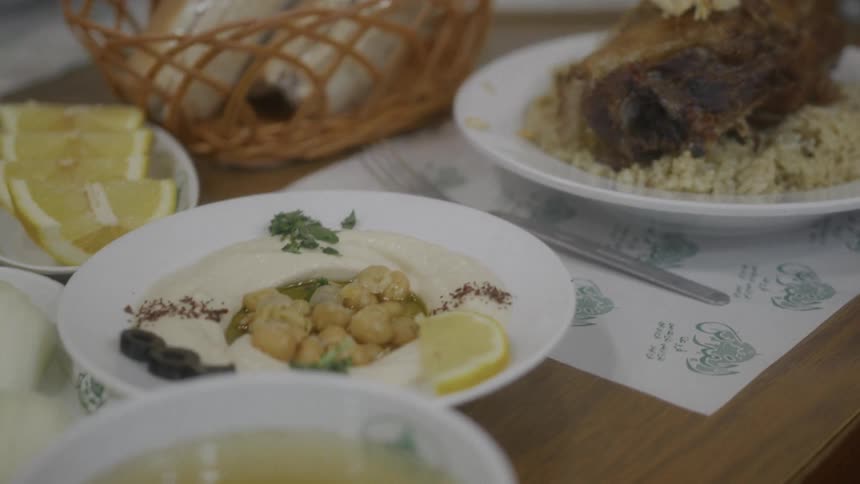 A slow rack focus shot reveals a traditional Middle Eastern meal of roasted lamb on rice, hummus with chickpeas, soup, and fresh pita bread