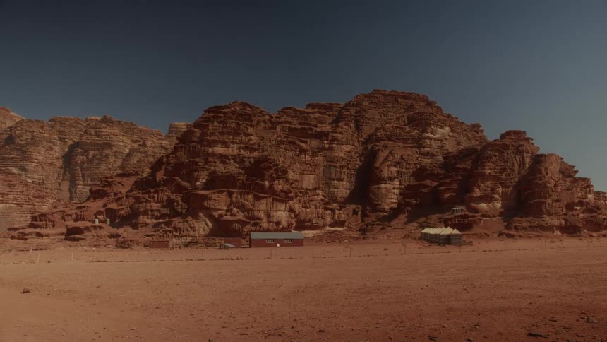 A remote settlement sits at the base of majestic red rock mountains under a clear blue sky in a vast desert landscape