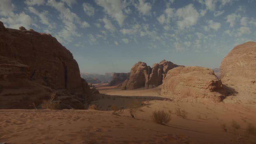 A cinematic wide shot of a red sand desert canyon with massive rock formations under a partly cloudy blue sky