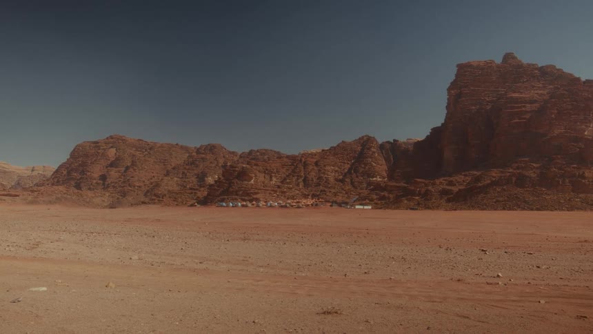 A wide shot of a small encampment at the base of towering red rock mountains in a vast, arid desert