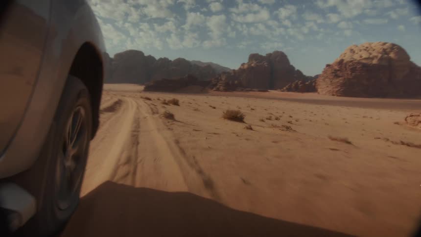 A thrilling point-of-view shot captures an SUV speeding across the vast, sandy desert, kicking up sand against a backdrop of majestic rock formations