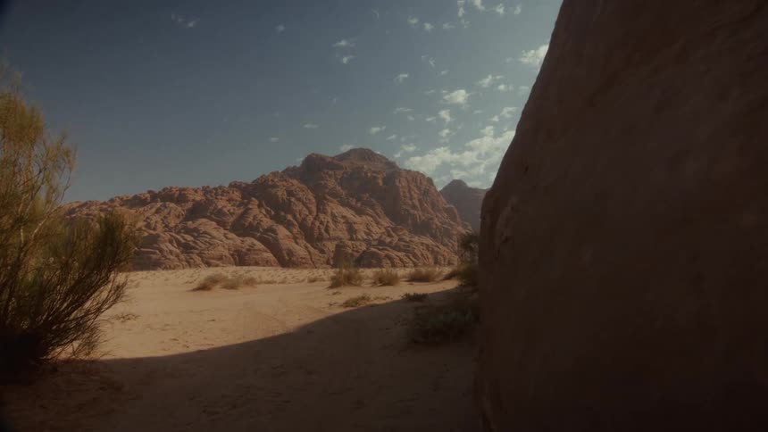 A slow pan reveals a sun-drenched desert landscape with rugged, rocky mountains under a clear blue sky