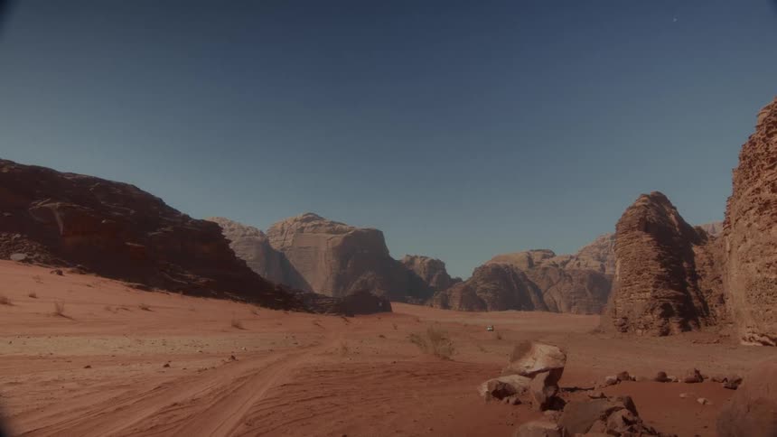 A lone vehicle drives through a vast red desert landscape with towering rock formations under a clear blue sky