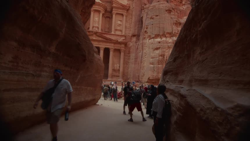 Slow tilt down reveals the magnificent Al-Khazneh Treasury at Petra through the narrow Siq canyon, as tourists gather to admire the ancient wonder