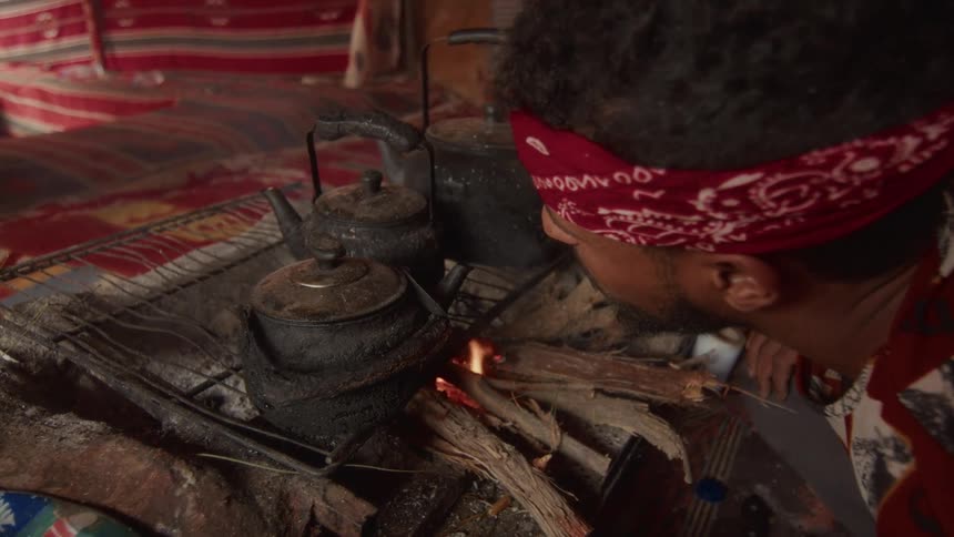 A man blows on the embers of an indoor fire to heat water in traditional blackened kettles