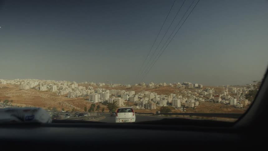 A point-of-view shot from inside a moving car driving towards a sprawling city built on arid hills