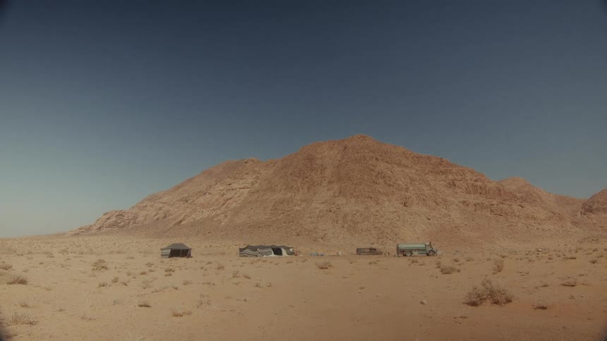 A wide, static shot reveals a desolate Bedouin camp with a water truck at the base of a large mountain in the desert