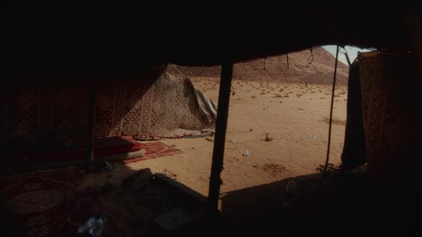 A view from inside a dark Bedouin tent looking out into the bright, windy desert with a pyramid in the distance