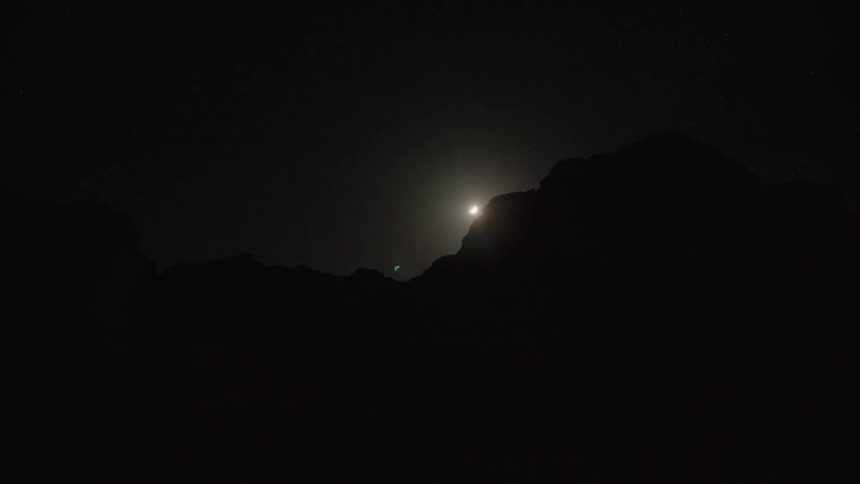 A time-lapse of the bright moon setting behind a silhouetted mountain range on a clear, starry night