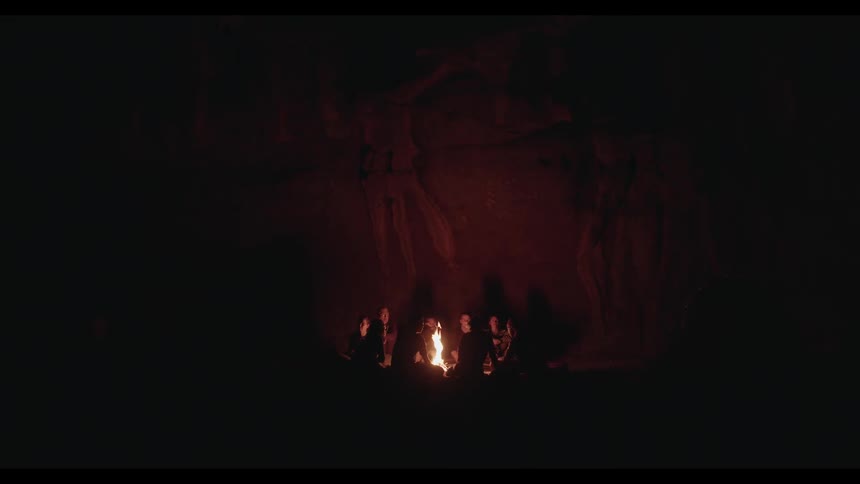 A group of people sit around a glowing campfire in a dark cave at night, their shadows dancing on the rock walls