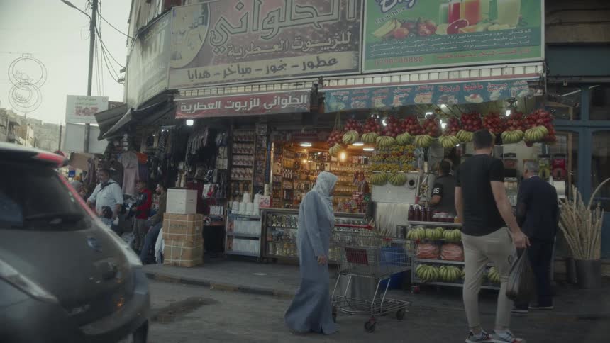 A car moves past revealing a bustling street corner market with pedestrians and shoppers in a Middle Eastern city