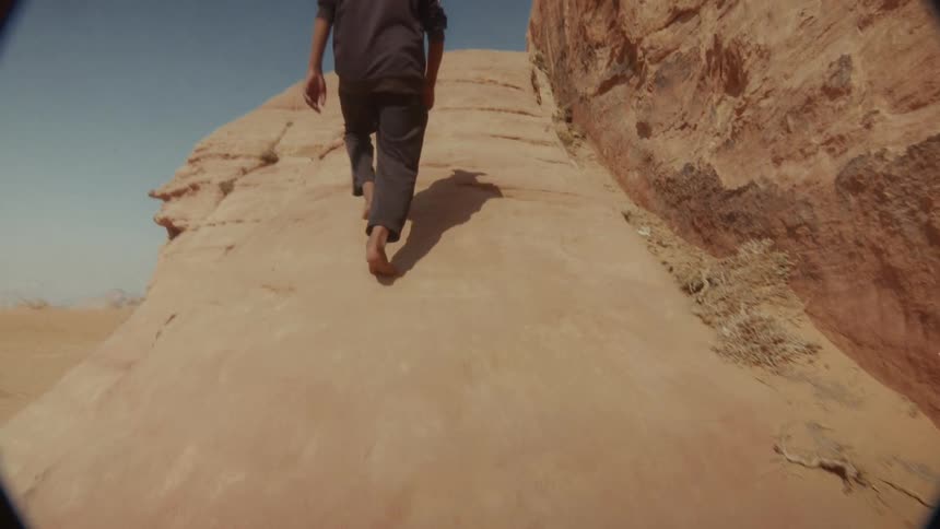 A barefoot young man carefully climbs a steep, sandy rock formation in the desert in slow motion