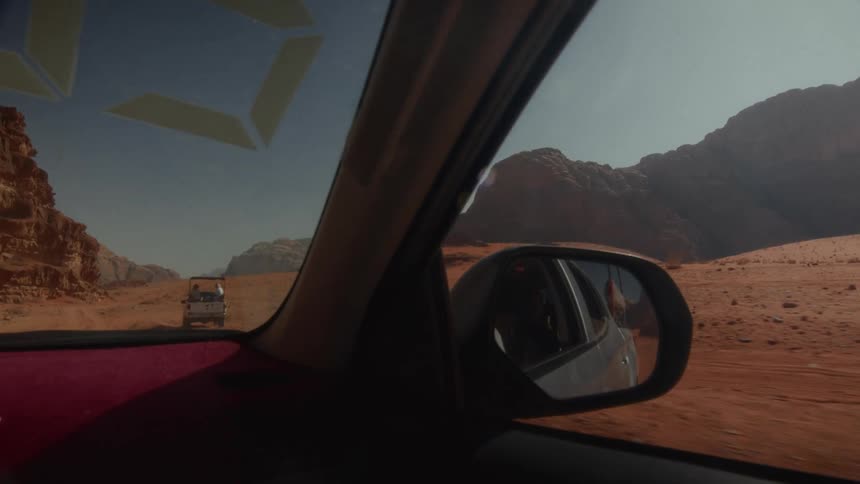 A point-of-view shot from a car driving through the Wadi Rum desert in Jordan, following another vehicle on a dusty trail