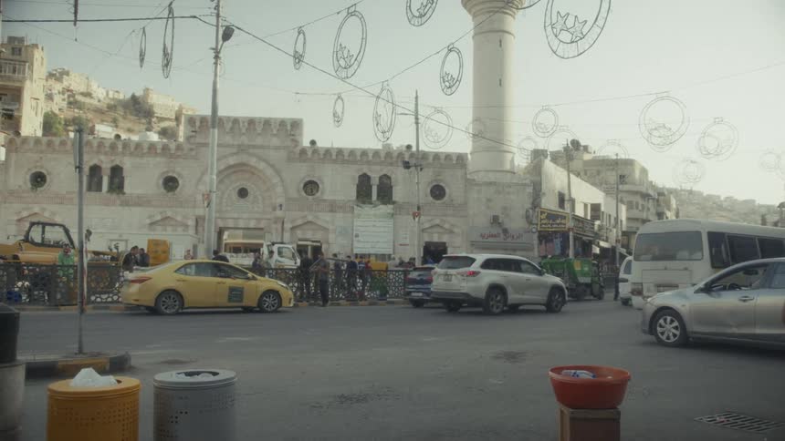 Traffic and pedestrians move through a busy street corner in a Middle Eastern city with a large mosque in the background