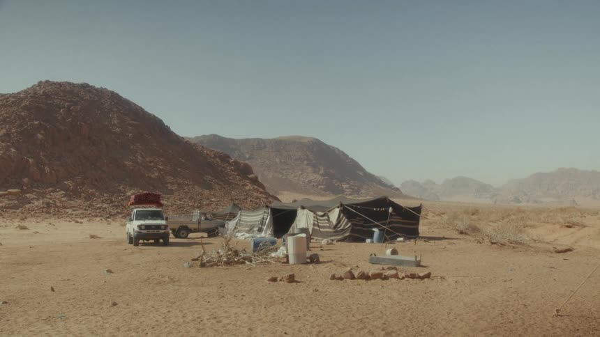 A wide, static shot of a Bedouin camp with trucks and tents in the vast desert landscape of Wadi Rum, Jordan