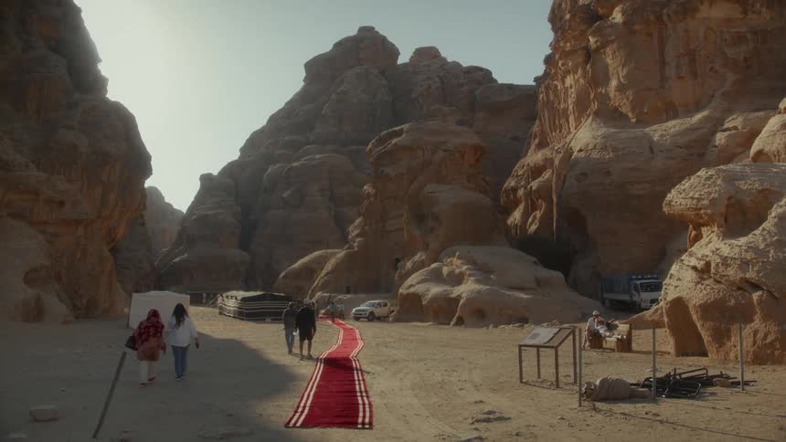 A long red carpet is rolled out through a sandy canyon as people arrive at a desert encampment