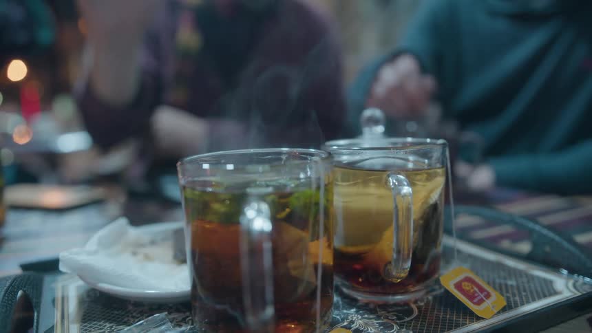 A person stirs and lifts a steaming glass of hot tea from a tray in slow motion at a cozy cafe
