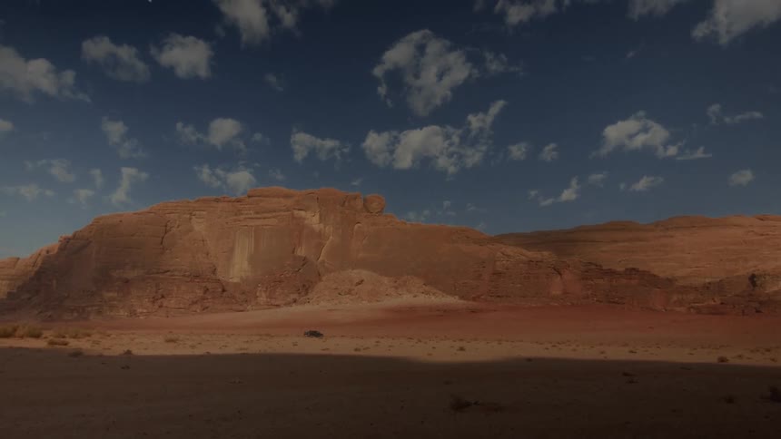 A timelapse of shadows moving across the vast red rock mountains of a desert landscape under a cloudy blue sky