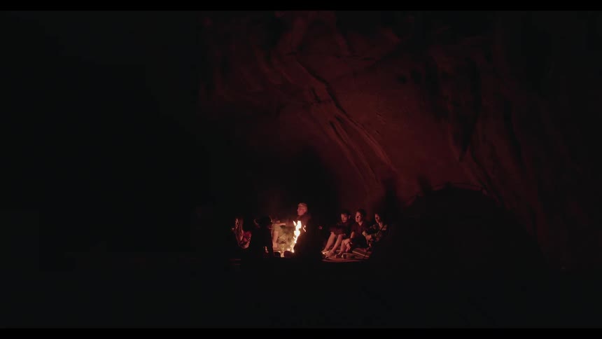 A group of friends huddle together around a warm campfire inside a dark, vast cave at night