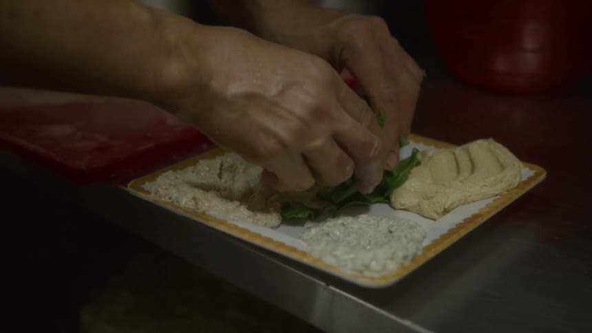 A chef carefully garnishes a plate of assorted Mediterranean appetizers with fresh greens in a restaurant kitchen