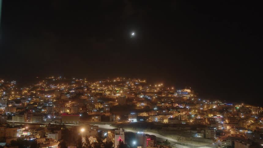A crescent moon hangs in the dark night sky over a brightly illuminated city built into a hillside, with cars driving on the roads below