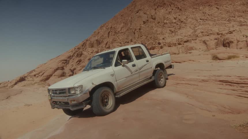 A white off-road pickup truck carefully navigates a steep rocky incline in a vast desert landscape under the hot sun