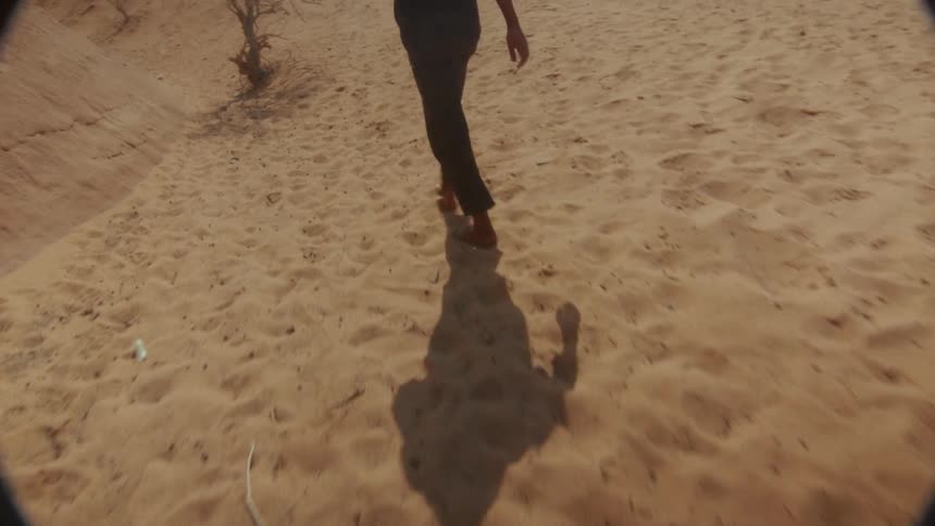 Slow-motion following shot of a man walking barefoot through a vast, hot desert landscape