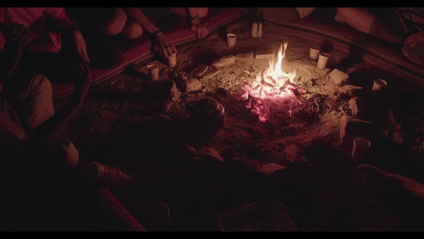 An overhead shot captures a group of friends sitting on a blanket around a crackling campfire at night, sharing stories and drinks