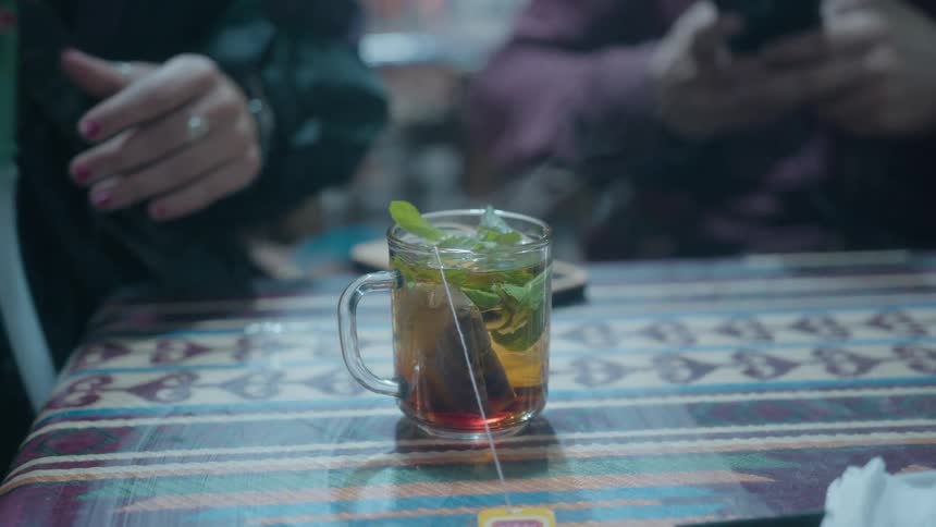 A hand adds fresh mint leaves to a steaming glass mug of hot tea on a patterned tablecloth