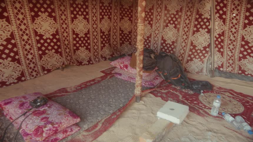 A slow tilt-down shot reveals the rustic interior of a traditional desert tent, complete with patterned fabric walls, colorful rugs on the sand, and a central fire pit
