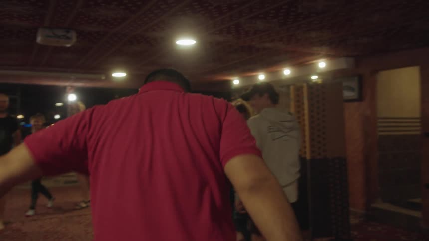 A man in a red shirt leads a group of people in a traditional folk dance at a party in a warmly lit room at night in slow motion