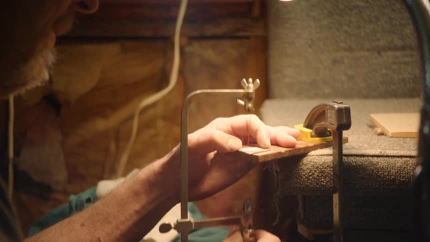 A skilled woodworker meticulously uses a coping saw to make an intricate cut on a small piece of wood in his workshop