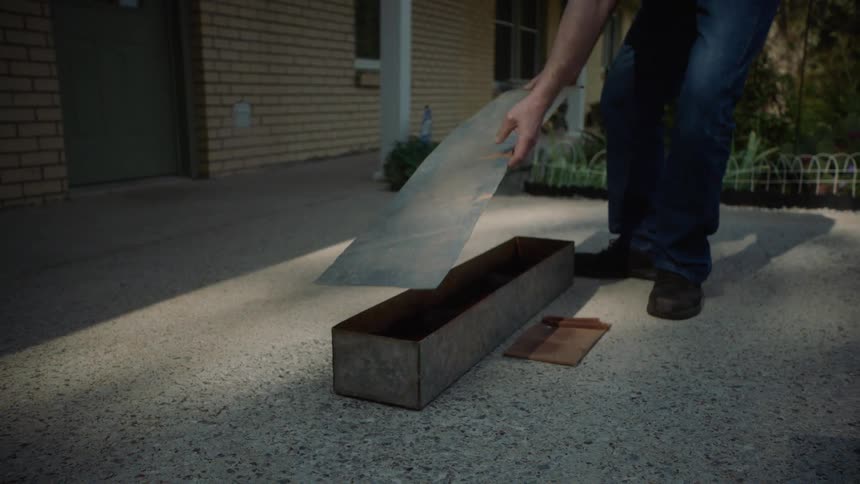 A man carefully places a sheet metal lid over a long metal container on a concrete driveway in slow motion