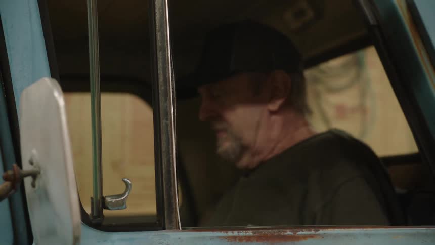 An older man in a baseball cap sits thoughtfully in the driver's seat of his vintage truck in a garage