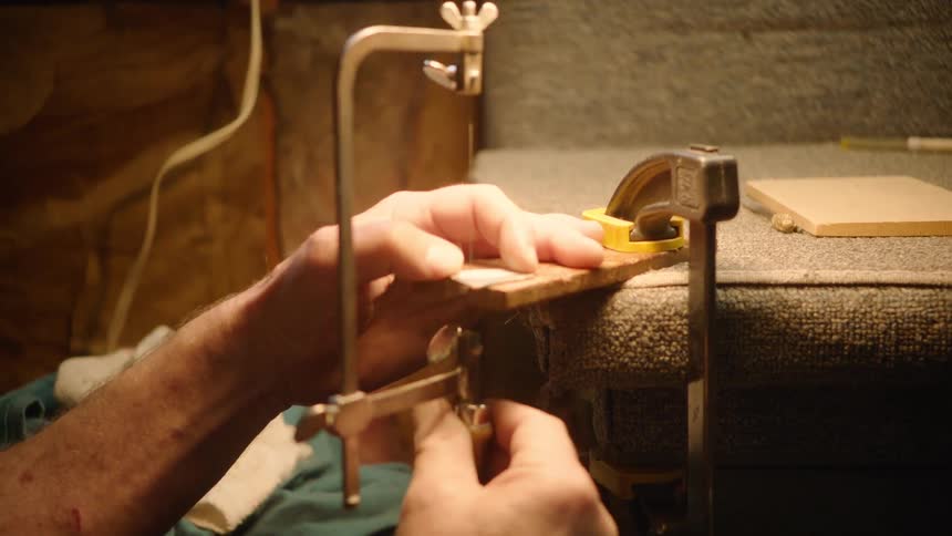 A craftsman carefully uses a coping saw to cut a detailed shape into a small piece of wood in his workshop