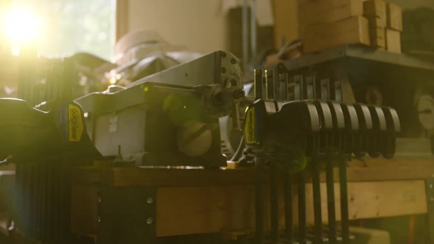 A slow pan reveals various woodworking clamps and machinery on a workbench in a sun-drenched, dusty workshop