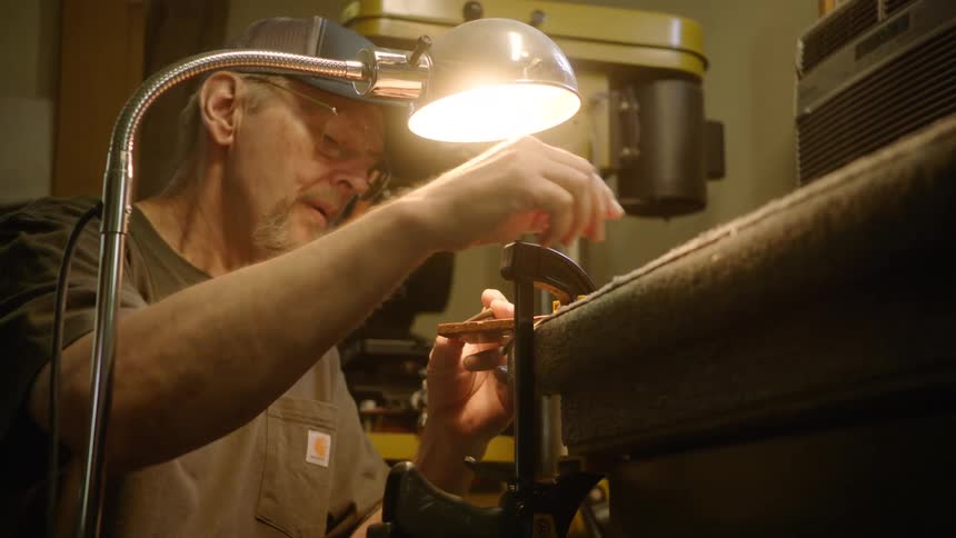 A focused artisan carefully sands a small wooden part held in a clamp under a task light in his workshop
