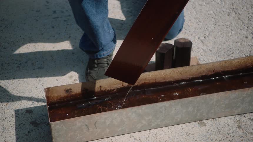 In super slow motion, a worker carefully pulls a long metal flashing out of a trough of dark, oily liquid, letting the excess drip off